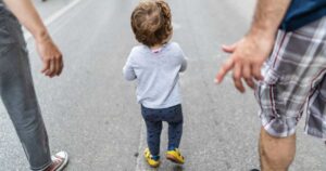 Toddler walking on city street with two adults nearby